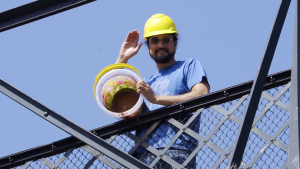 A still from The Pee Pee Poo Poo Man, showing the titular character donning a construction helmet and waving while holding a turned over bucket lined with a brown substance.